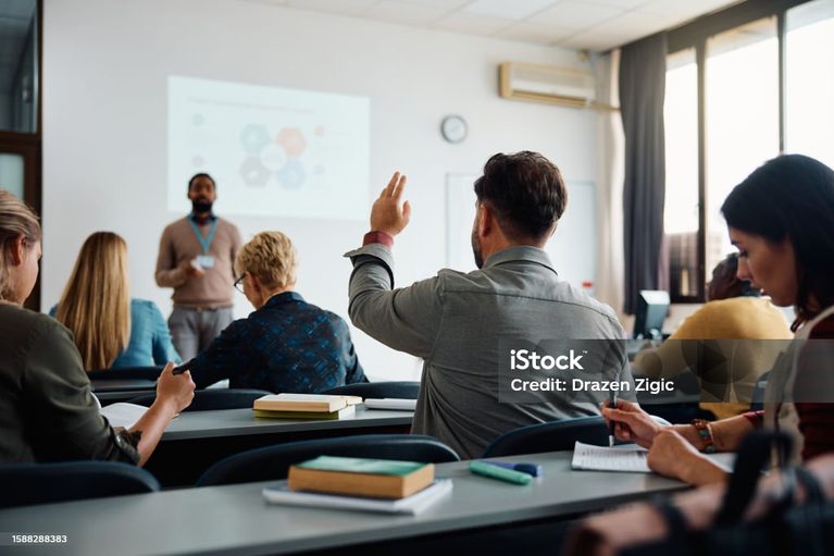 Rear view of man raising arm to ask a question during a presentation in lecture hall.